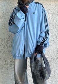Personne portant une veste Adidas bleu clair surdimensionnée avec des rayures noires, un pantalon gris, des gants noirs sans doigts, tenant un sac noir et gris à pois.