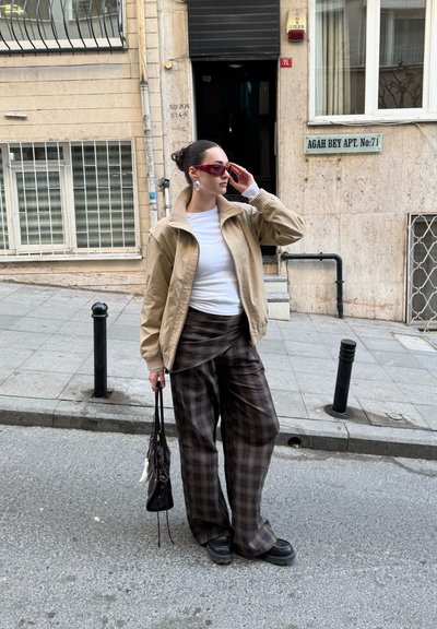Mujer con chaqueta beige y pantalones anchos de cuadros de pie en la calle de la ciudad sosteniendo una bolsa negra, ajustándose unas gafas de sol rojas.