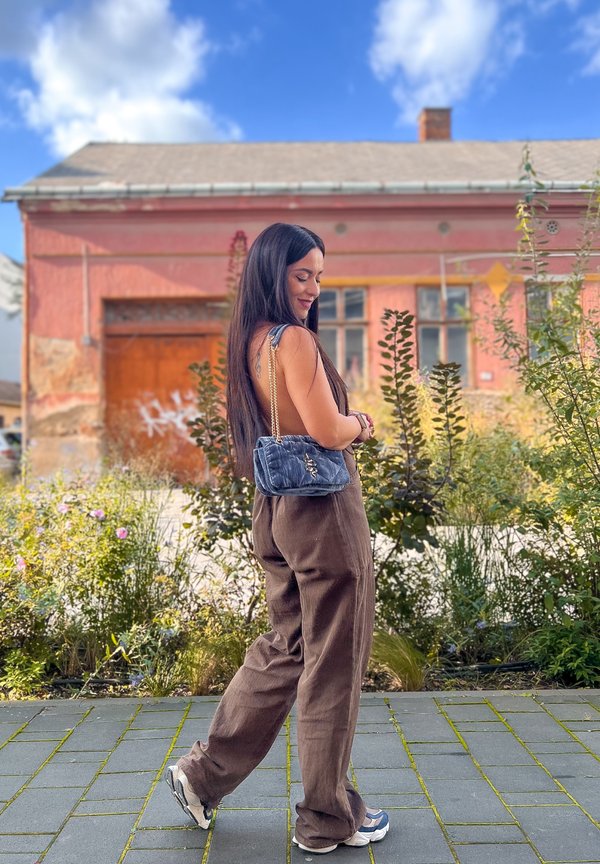 Donna con capelli lunghi, in pantaloni marroni larghi e sneaker, che porta una borsa a spalla blu, in piedi su un sentiero pavimentato con piante e un edificio rosso sullo sfondo.
