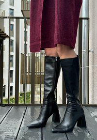 Μαύρες δερμάτινες μπότες μέχρι το γόνατο με αιχμηρή μύτη και χοντρό τακούνι; διαθέτουν πλαϊνό φερμουάρ και λεία υφή. Φοριούνται με ένα μπορντό πλεκτό φόρεμα.