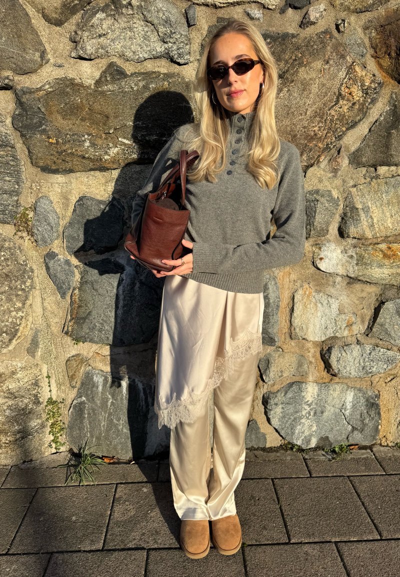 Pull en tricot gris avec détail de boutons, pantalon en satin crème avec bordure en dentelle, et chaussons en daim marron, assortis à un sac à bandoulière en cuir marron.