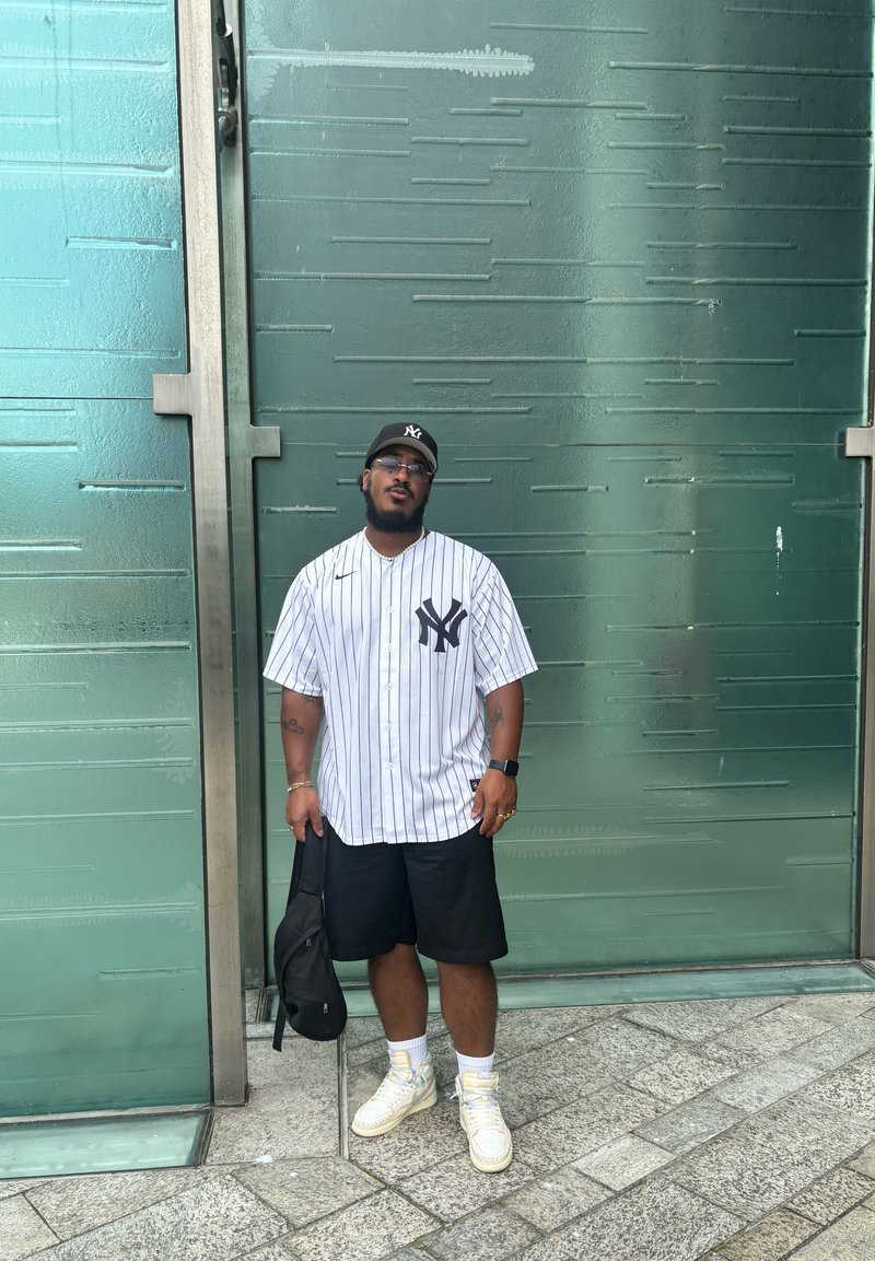 Μπλουζάκι μπέιζμπολ με μαύρες και λευκές ρίγες και λογότυπο NY, μαύρες βερμούδες, λευκά αθλητικά παπούτσια, κρατώντας μια μαύρη τσάντα, στέκεται μπροστά από ένα υφασμάτινο πράσινο φόντο.