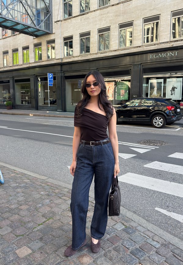 Kvinde med langt mørkt hår, solbriller, off-shoulder top og brede jeans står på en bygade og holder en taske nær et fodgængerfelt.