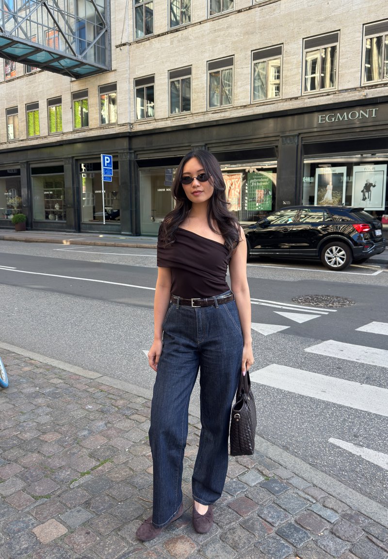 Femme aux longs cheveux foncés, portant des lunettes de soleil, un haut épaules nues et un jean large, debout dans une rue de la ville, tenant un sac à main près d’un passage piéton.
