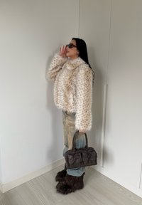Veste en fausse fourrure crème avec texture tachetée, jeans amples, lunettes de soleil noires surdimensionnées, et sac à main en cuir foncé avec accents de nœud.