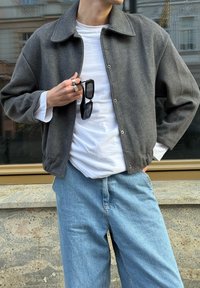 Grau gerippte Jacke mit Kragen, Knopfverschluss und elastischem Saum. Kombiniert mit einem weißen Hemd und hellblauen Jeans. Zubehör umfasst eine Sonnenbrille.