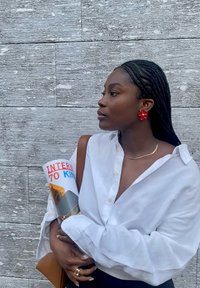 Une personne portant une chemise blanche à boutons tient un magazine roulé avec des textes rouges et bleus. De remarquables boucles d'oreilles en forme de fleurs rouges et une chaîne en or sont visibles.
