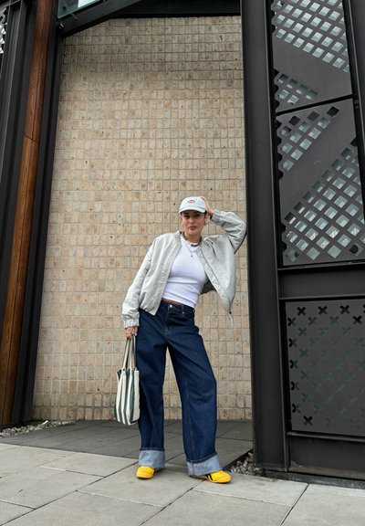 Chaqueta bomber gris claro, top corto blanco, jeans de pierna ancha azules, zapatos amarillos de lona, bolsa de rayas, de pie contra una pared texturizada.