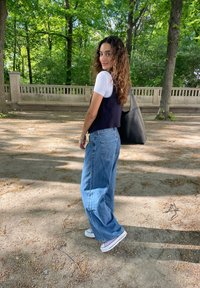 Μοντέλο φορώντας ένα εφαρμοστό ναυτικό γιλέκο πάνω από ένα λευκό μπλουζάκι, χαλαρές μπλε τζιν και μαύ�ρα αθλητικά παπούτσια, κρατώντας μια μαύρη τσάντα, σε μια δασώδη περιοχή.