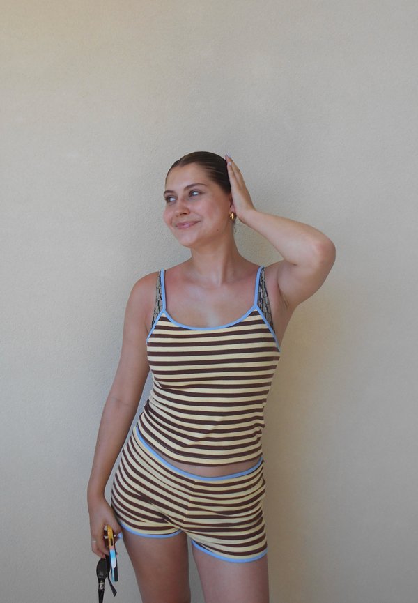 Jeune femme en débardeur rayé marron et beige et short, souriante et touchant sa tête, tenant des lunettes de soleil et un téléphone contre un mur uni.