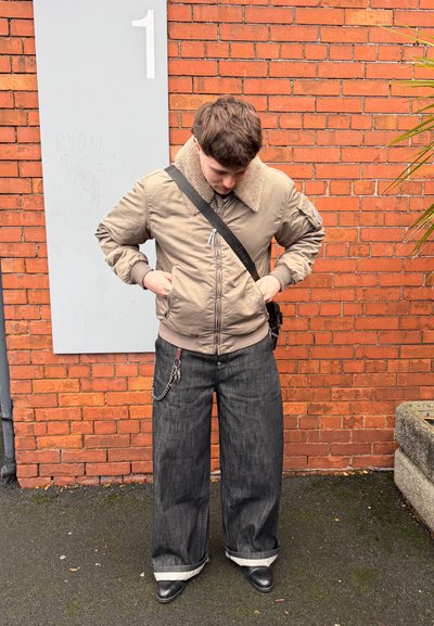 Joven ajustando los bolsillos de una chaqueta beige de piel vuelta, vestido con jeans anchos oscuros y zapatos negros, de pie contra una pared de ladrillos naranja junto a un cartel gris.