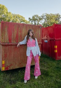 Gilet rosa a fantasia sopra una camicia azzurra chiaro, abbinato a pantaloni rosa a zampa di elefante. L'outfit è completato da scarpe bianche su uno sfondo arrugginito.