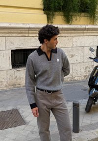 Pull-over tricot gris avec col noir, associé à un pantalon beige. Présente un tissu texturé et un petit logo sur la poitrine. Vue de profil.