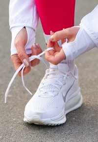 Chaussure de course blanche avec dessus en mesh, accents gris et lacet blanc. Des mains ajustent les lacets, posée sur une surface grise.