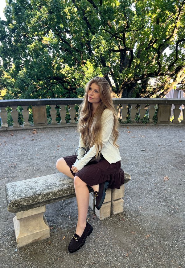 Jeune femme aux longs cheveux blonds portant un pull blanc et une jupe foncée, assise sur un banc en pierre dans un parc avec de grands arbres feuillus.