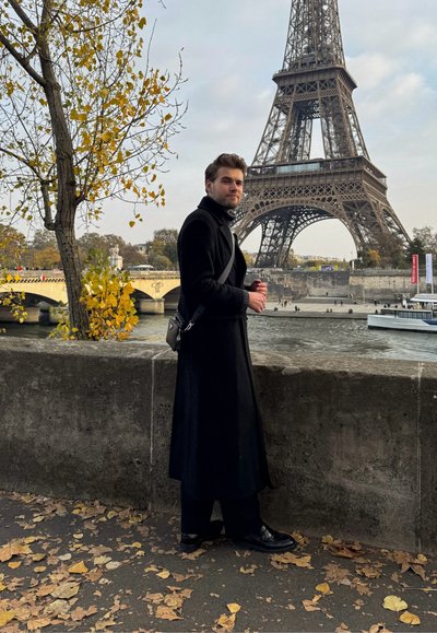 Una persona con un largo abrigo negro está de pie junto a una pared de piedra cubierta de hojas amarillas, con la Torre Eiffel visible al fondo.