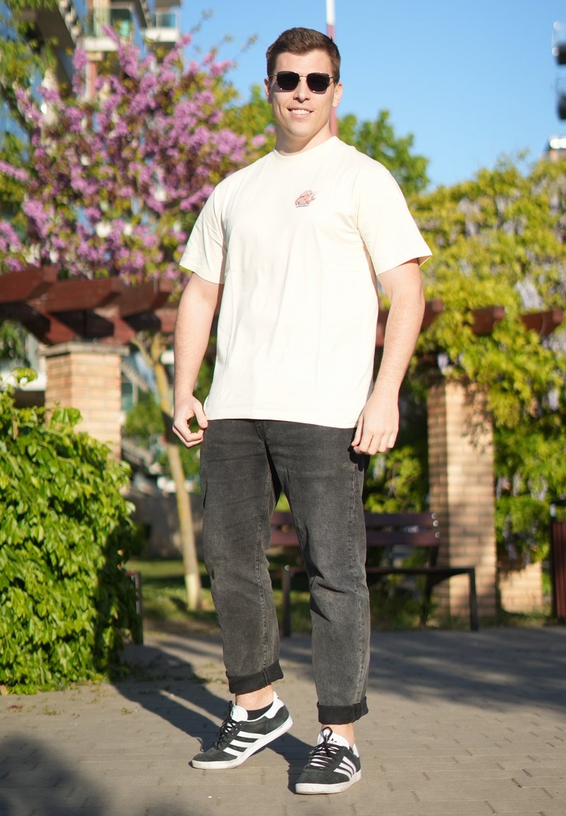 Jeune homme portant des lunettes de soleil, un t-shirt crème, un jean noir et des baskets, debout en extérieur par une journée ensoleillée avec des arbres et des bancs derrière lui.