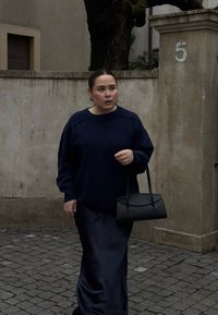 Navyblauwe gebreide trui met vallen schouders, gecombineerd met een lange donkere satijnen rok. Houdt een kleine zwarte handtas vast en staat op straatstenen.