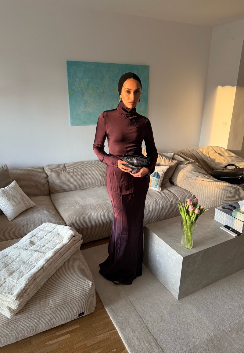 Femme en robe à manches longues violet foncé et foulard, debout dans un salon baigné de soleil, tenant une pochette noire, près d'un canapé et d'une table basse en marbre.