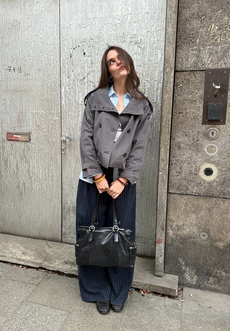 Fiatal nő szürke zakóban és csíkos nadrágban, fekete kézitáskát tartva, egy városi járdán áll, egy texturált fémajtó előtt.