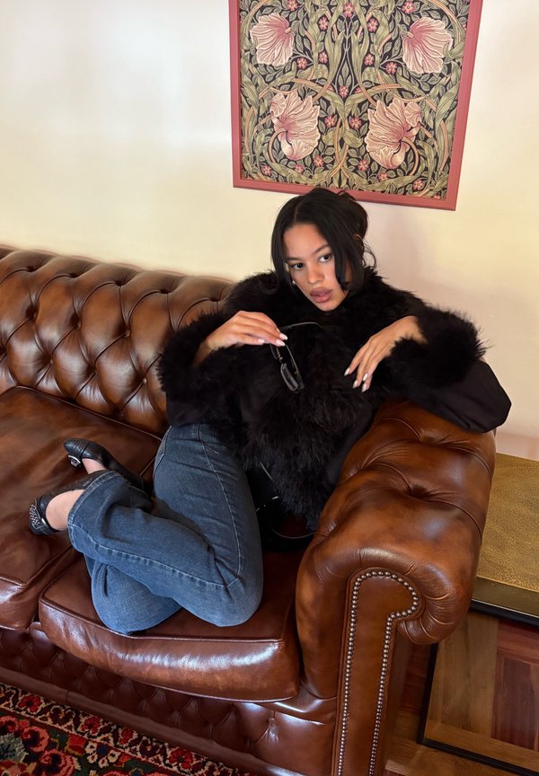 Un manteau en fausse fourrure noir, un jean en denim foncé et des chaussures noires sur un canapé en cuir marron. En arrière-plan, une œuvre d'art murale florale.