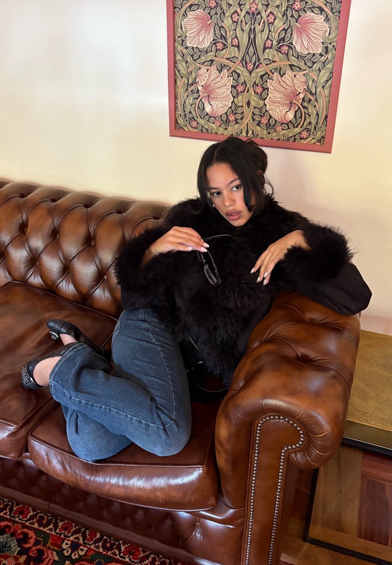 Czarny płaszcz z futerkiem, ciemnoniebieskie dżinsy i czarne buty na brązowej skórzanej sofie. Tło zdobi kwiatowy obraz.