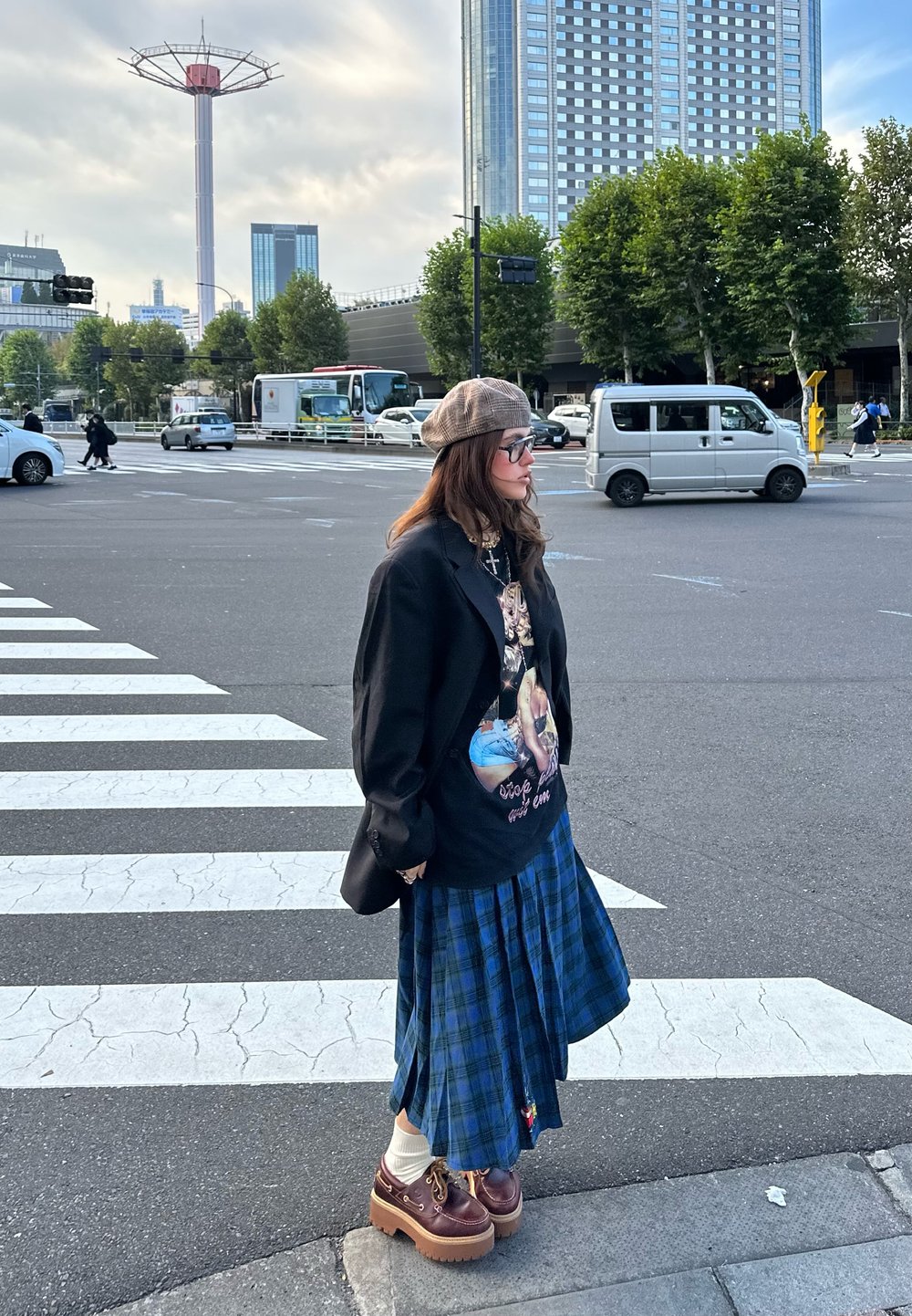 Czarna, oversize'owa marynarka, T-shirt z grafiką, niebieska spódnica w kratę i brązowe buty na platformie w białych skarpetkach. Osoba stoi na przejściu dla pieszych.