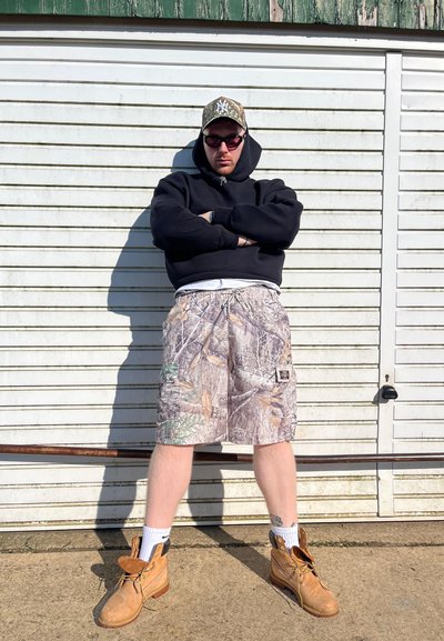 Sudadera negra, pantalones cortos de camuflaje con un patrón de hojas, botas marrones, calcetines blancos. Lleva gafas de sol y una gorra de camuflaje, de pie contra un cobertizo blanco.