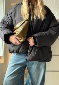 Casaco puffer preto com gola alta, combinado com jeans de perna larga azuis claros. Segurando uma mala de mão texturizada em verde-azeitona.