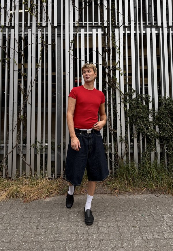 Person med kort blondt hår iført en rød skjorte, sorte vide shorts, hvide sokker og sorte sko står ved et højt metalhegn med klatreplanter.