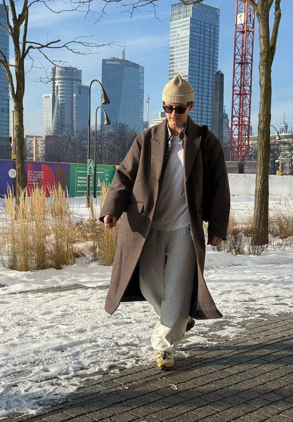 Junger Mann mit Sonnenbrille, beiger Mütze, langem kariertem Mantel und grauen Jogginghosen geht auf einem schneebedeckten Bürgersteig in einem urbanen Gebiet mit hohen Gebäuden.