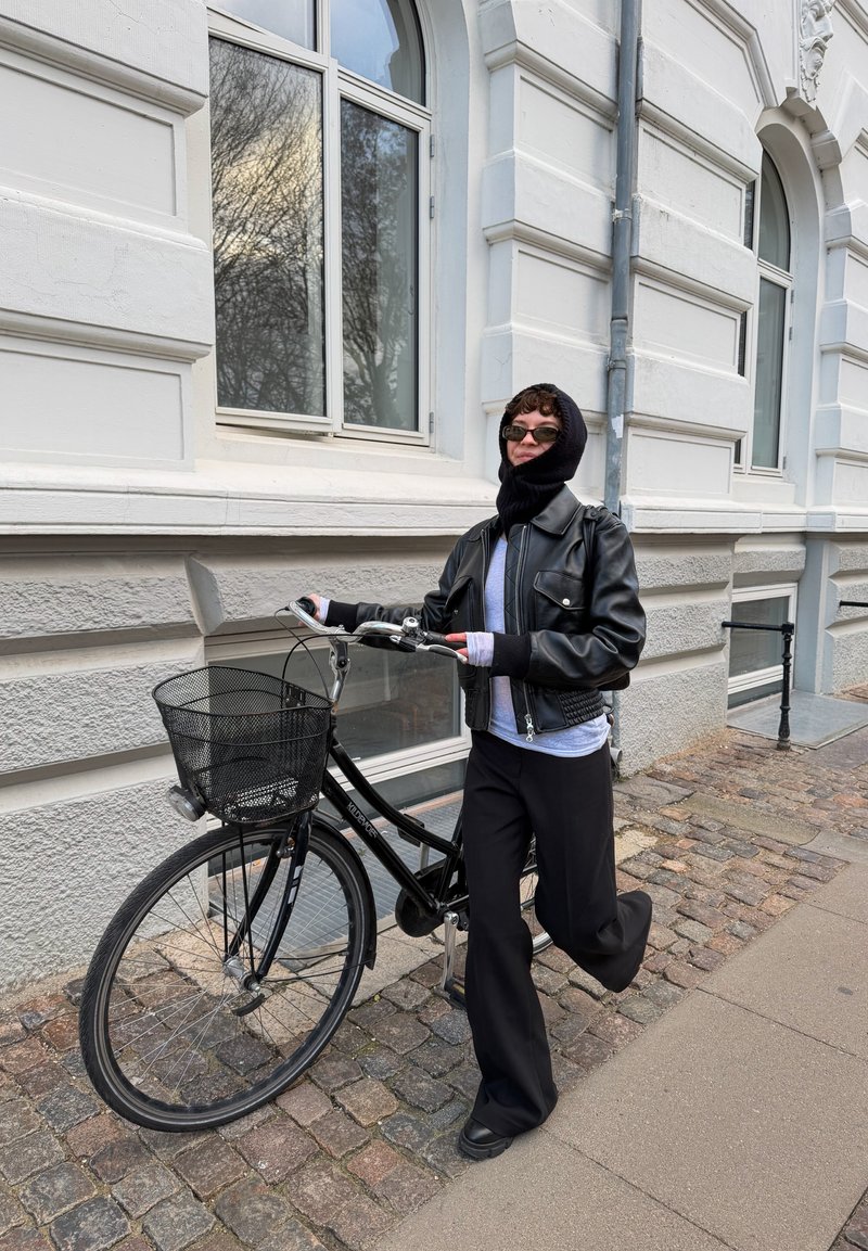 Black bicycle with a mesh basket on cobblestone street; the person wears a black leather jacket, gray shirt, and black pants.
