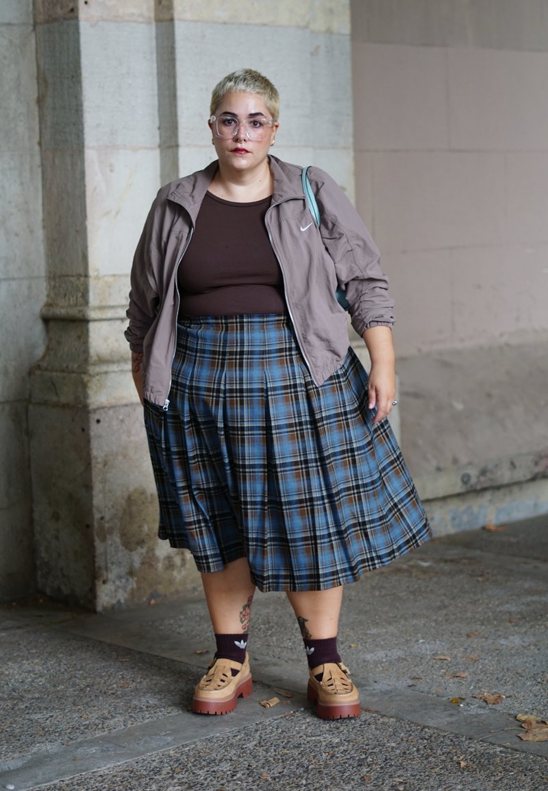 Haut marron à manches courtes, veste zippée beige, jupe plissée à carreaux bleus et noirs, chaussures beiges à semelles épaisses, chaussettes bordeaux.