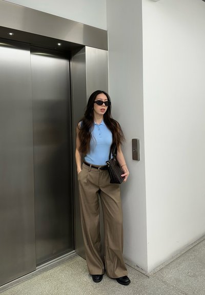 Mujer con cabello largo, gafas de sol, camiseta azul sin mangas y pantalones beige de pierna ancha, de pie cerca de puertas de ascensor de acero inoxidable cerradas.