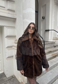 Rudos spalvos dirbtinio kailio paltas su plačiu apykakliu, turintis dviejų tonų dizainą. Modelis dėvi apvalias saulės akinių ir laiko mažą rankinę.