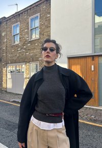 Svart oversized frakk over en grå rullekrage, beige bukser med høy midje og et rødt belte. Tilbehør inkluderer runde mørke solbriller og store hoop-øredobber.