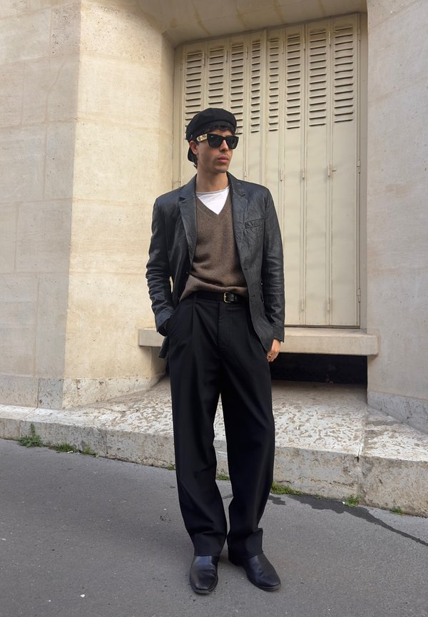 Man wearing black beret, sunglasses, leather jacket, brown jumper, wide black trousers, and black shoes standing against beige stone wall.