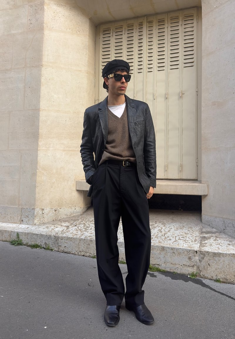Homme portant un béret noir, des lunettes de soleil, un blazer en cuir, un pull marron, un pantalon large noir et des chaussures noires, debout contre un mur en pierre beige.
