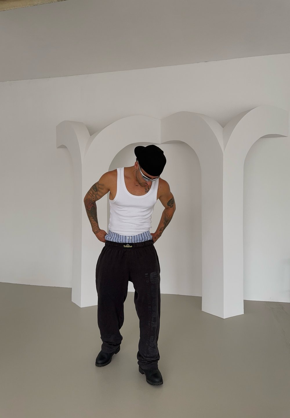 Top branco sem mangas, calças de treino pretas com detalhe gráfico, botas pretas e um boné preto. Com fundo minimalista branco com arcos.