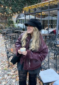 Maroon shearlingjakke med struktureret overflade, lagdelt over en grå top og en stribet sort nederdel. Holder en lille kop kaffe.