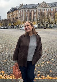 Giacca marrone oversize sopra un maglione grigio, abbinata a jeans scuri. Tiene in mano una borsa intrecciata color corallo. Strada acciottolata con foglie d'autunno.