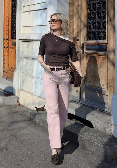 Mujer con cabello corto y rubio, usando gafas grandes, una blusa negra asimétrica, pantalones rosas y mocasines marrones, caminando junto a una puerta de madera.