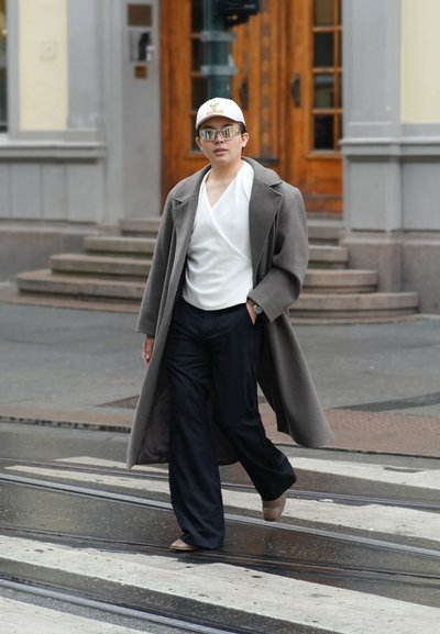 Persona con abrigo gris de gran tamaño, blusa blanca, pantalones negros, zapatos beige, gorra blanca con caricatura y gafas de sol reflectantes caminando por un paso de peatones en la ciudad.