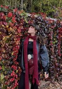 Černá kožená bunda, burgundský pletený šátek, přiléhavý černý top, sukně s vysokým pasem a ozdobným opaskem, strukturované legíny, bujný podzimní listnatý pozadí.