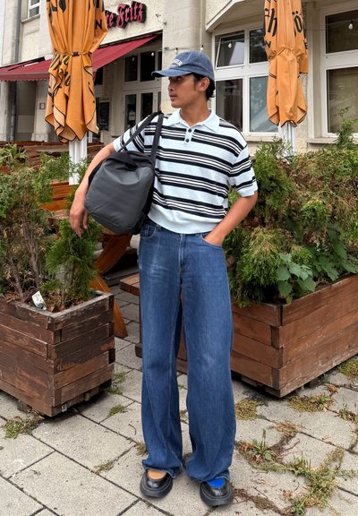 Joven con polo a rayas, jeans anchos y zapatos desiguales está de pie fuera de un café, sosteniendo una bolsa negra sobre el hombro.