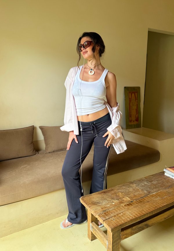 Junge Frau mit Sonnenbrille, weißem Tanktop und dunkler Hose posiert lässig neben einem beigen Sofa und einem rustikalen Holztisch im Innenbereich.