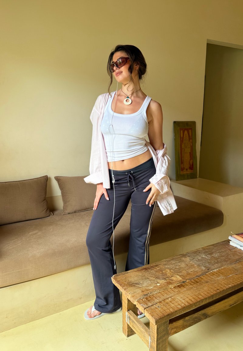 Jeune femme portant des lunettes de soleil, un débardeur blanc et un pantalon sombre, posant de manière décontractée à côté d'un canapé beige et d'une table en bois rustique à l'intérieur.