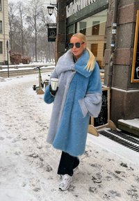 Μακρύ ψεύτικο παλτό από γούνα σε αποχρώσεις του μπλε και του γκρι, σε υπερμεγέθη σιλουέτα, με μεγάλο γιακά, μεγάλες κουμπότρυπες και τσέπες. Κρατώντας ένα φλυτζάνι καφέ.