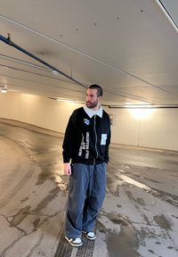 Μαύρο zip-up μπουφάν με λευκό γιακά και κείμενο, σε συνδυασμό με χαλαρούς γκρι παντελόνες και αθλητικά παπούτσια, standing σε ένα φωτεινό γκαράζ.