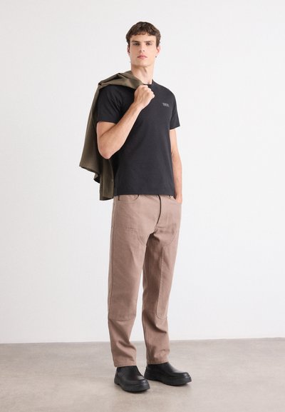 Camiseta negra, pantalones beige con costuras texturizadas y zapatos negros. Chaqueta verde oliva colgada sobre el hombro. Diseño minimalista.